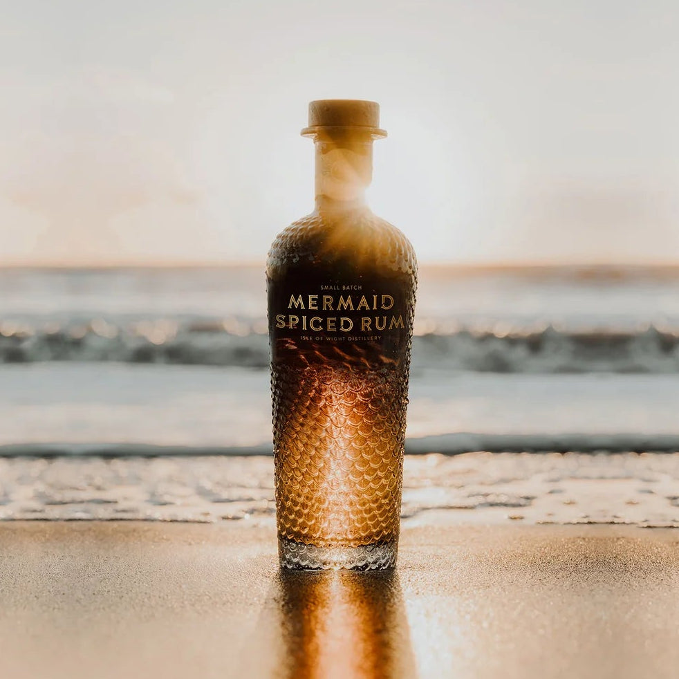 Load image into Gallery viewer, Mermaid Spiced Rum bottle on a sandy beach with a blurred sunset and ocean waves in the background