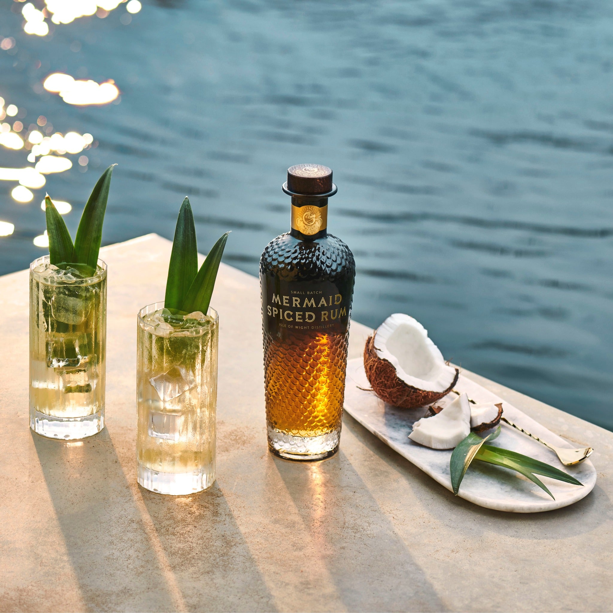 Load image into Gallery viewer, Mermaid Spiced Rum bottle with cocktails and coconut on a table by water