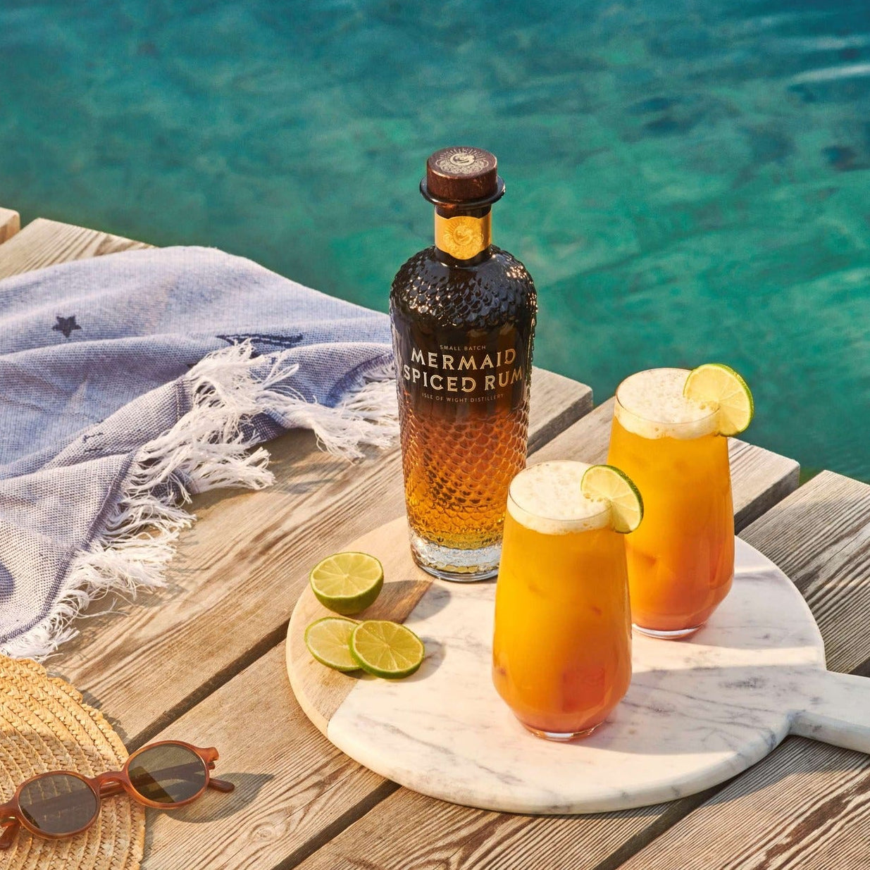 Load image into Gallery viewer, Bottle of Mermaid Spiced Rum with two glasses of rum punch on a wooden deck by a pool.