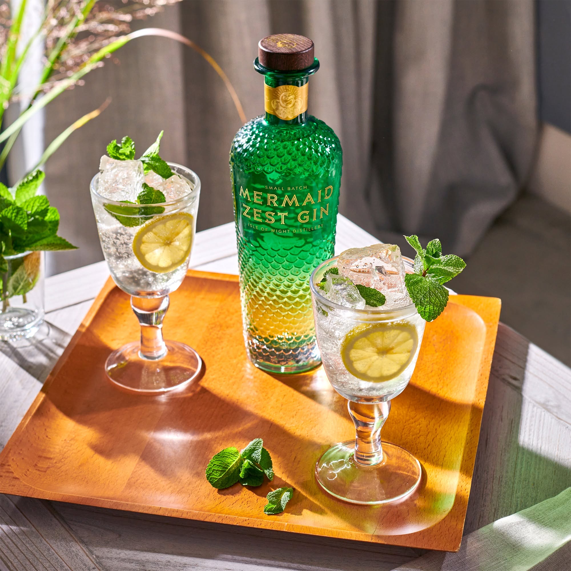 Load image into Gallery viewer, A bottle of Mermaid Zest Gin stands on a wooden tray beside two stemmed glasses filled with a clear cocktail, ice, mint leaves, and lime slices, set on a table with soft natural light and a curtain in the background.