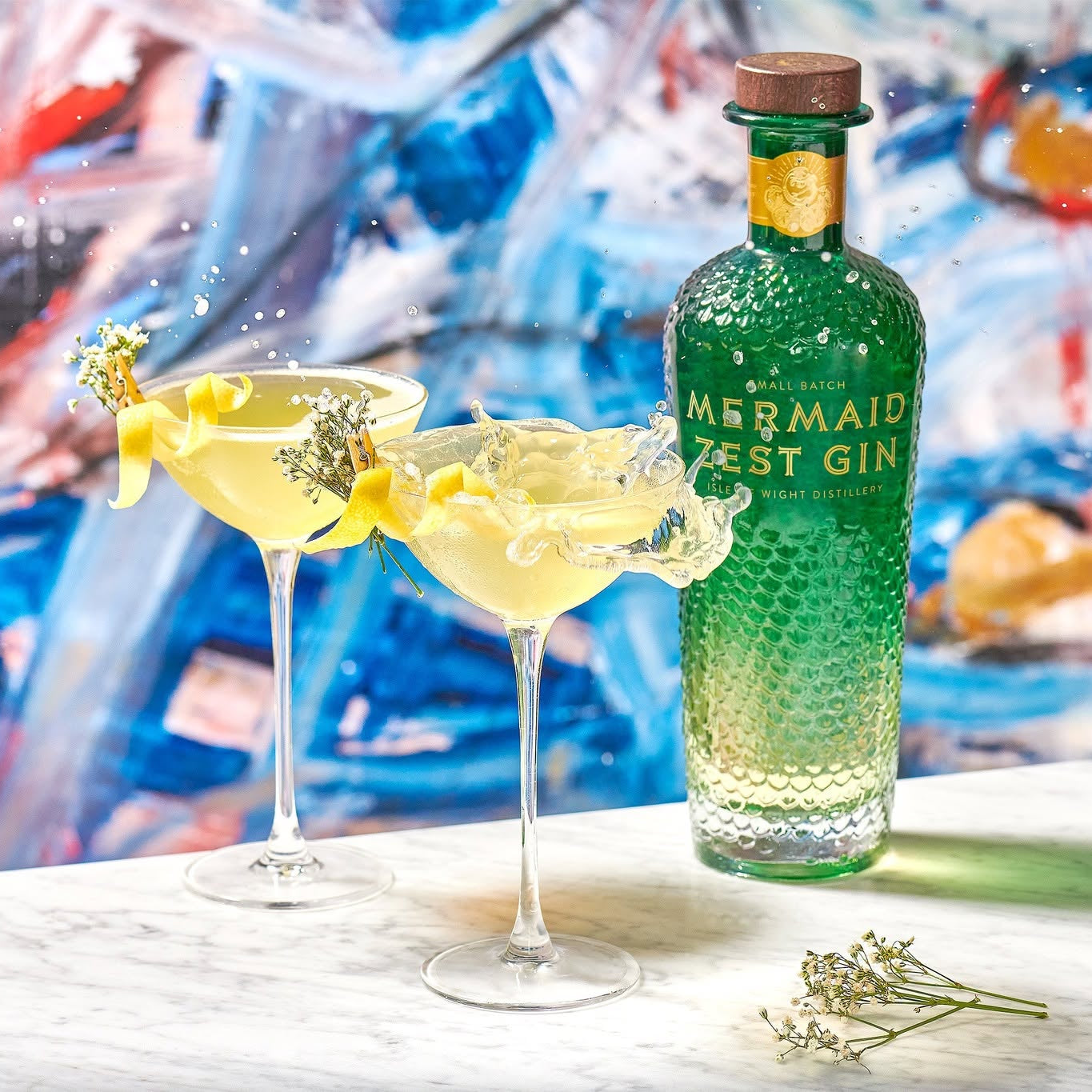 Load image into Gallery viewer, A green bottle of Mermaid Zest Gin stands on a white marble surface beside two margarita glasses filled with pale yellow drink and garnished with citrus and flowers, against a colourful abstract painted background.