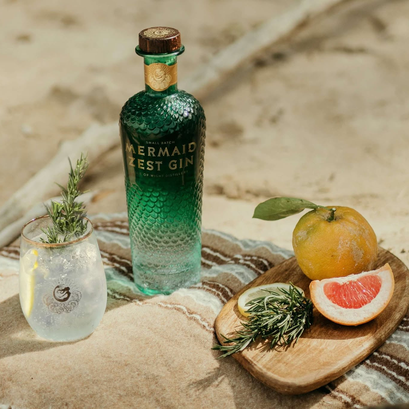Load image into Gallery viewer, A bottle of Mermaid Zest Gin stands on a beach blanket beside a glass of mixed drink, with a grapefruit, rosemary sprig, and wooden board in the sand.