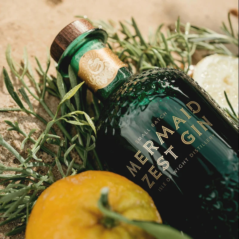Load image into Gallery viewer, Mermaid Zest Gin bottle with herbs and an orange on a sandy background