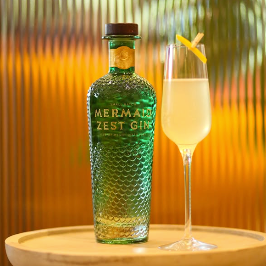 Load image into Gallery viewer, A bottle of Mermaid Zest Gin stands on a round wooden tray beside a filled coupe glass with a lemon twist, set against a warm orange and gold striped background.