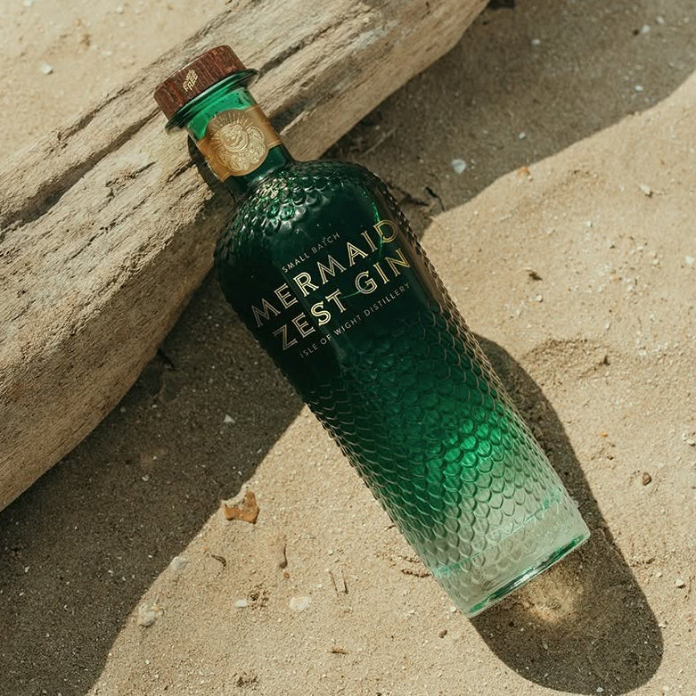 Load image into Gallery viewer, A green textured bottle of Mermaid Zest Gin lies on a sandy beach next to a piece of driftwood in bright natural sunlight.