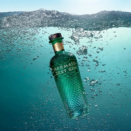 Load image into Gallery viewer, A green glass bottle labeled 'Mermaid Zest Gin' floating underwater in a ocean scene, with bubbles around it and four gold-toned seals aligned along the bottom.