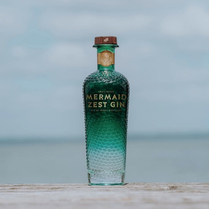 Load image into Gallery viewer, A bottle of Mermaid Zest Gin stands on a light surface with a blurred ocean and cloudy sky background.