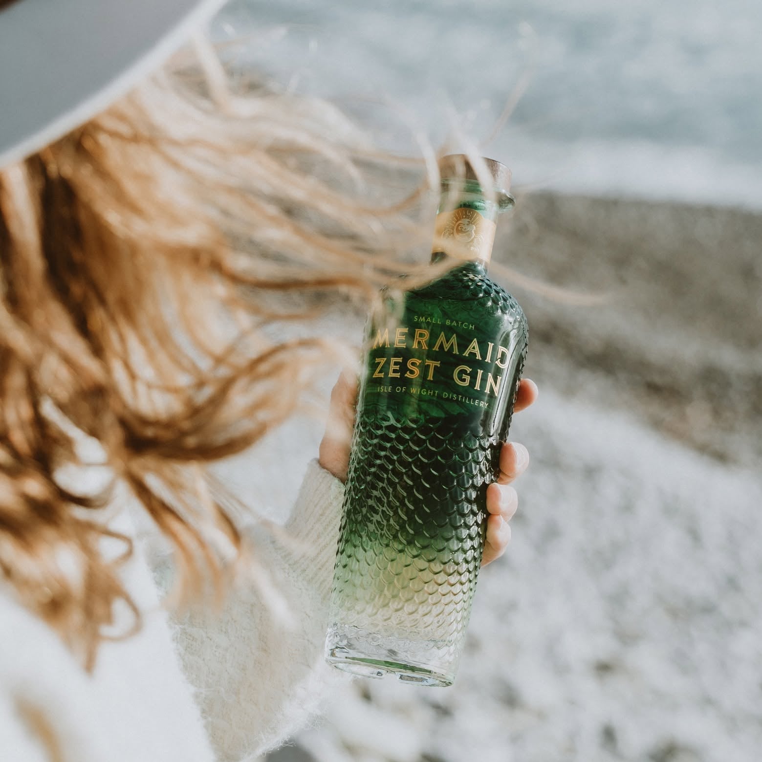 Load image into Gallery viewer, A woman on a beach holds a bottle of Mermaid Zest Gin. The person is wearing a white knit sweater and a white sun hat, with their blonde hair blowing in the wind.