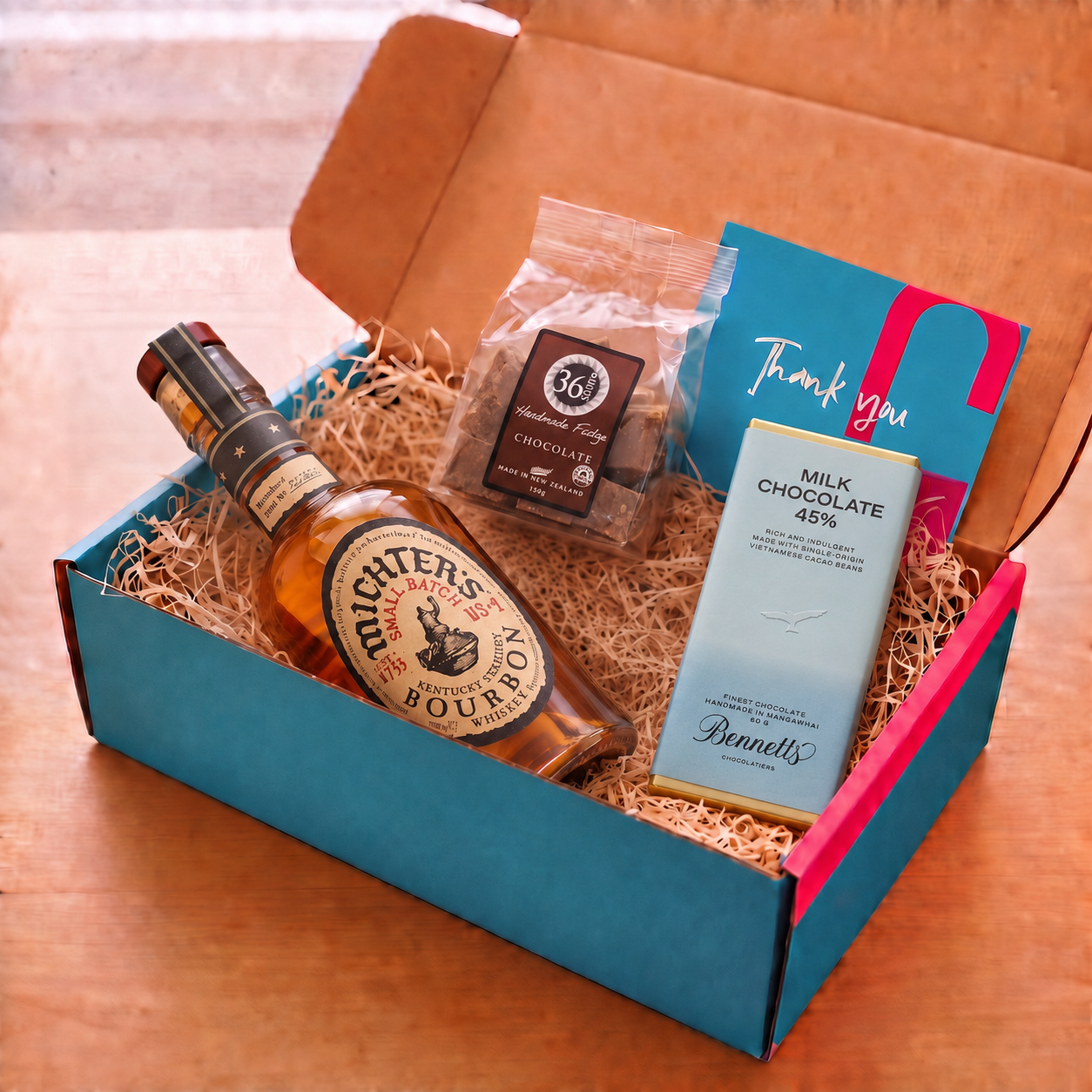 Load image into Gallery viewer, A curated gift box set on a wooden table, featuring a bottle of Michter’s US★1 Small Batch Kentucky Straight Bourbon, a 36 Degrees chocolate fudge packet, a Bennett's milk chocolate bar, and a blue and pink thank you card inside a COVE 27 cardboard box with wood-wool.
