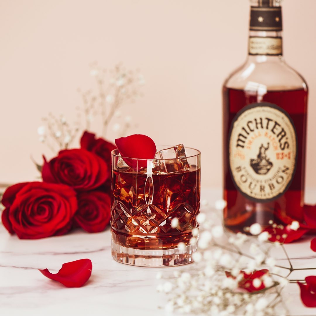 Load image into Gallery viewer, Glass of bourbon with ice and rose petal next to a bottle of Michter's bourbon and red roses.