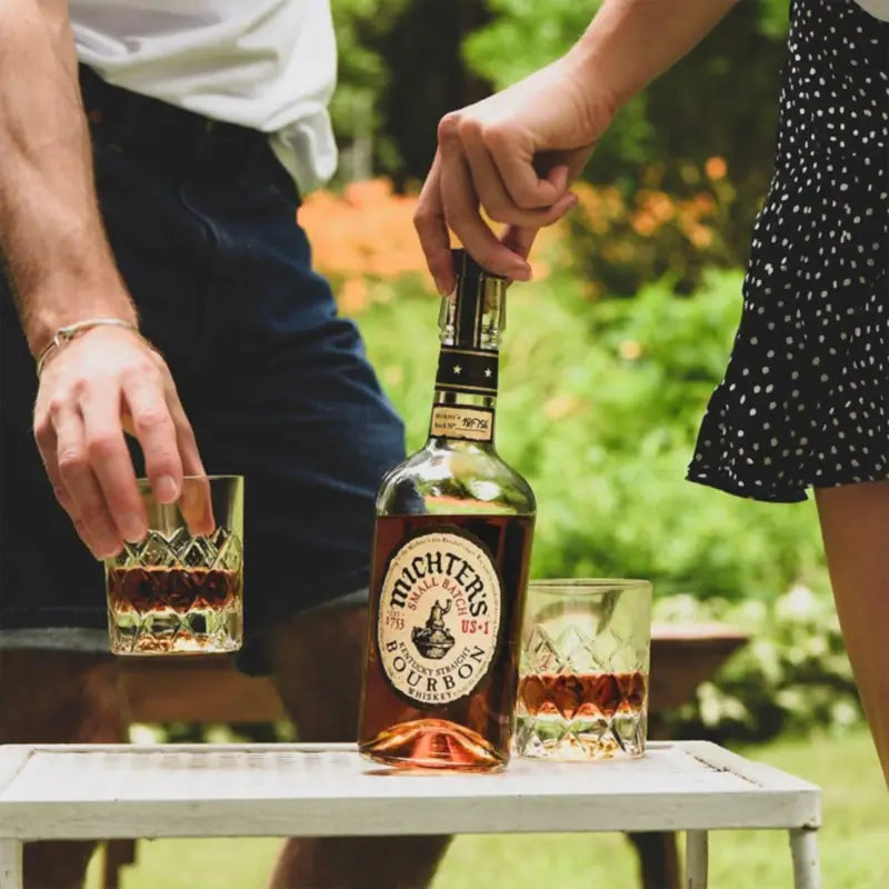 Load image into Gallery viewer, A bottle of Michter’s US1 Small Batch Kentucky Straight Bourbon whiskey is centred on a small white table outdoors, with two people partially visible holding or pouring near the bottle and two glasses. The background is a blurred green garden setting.