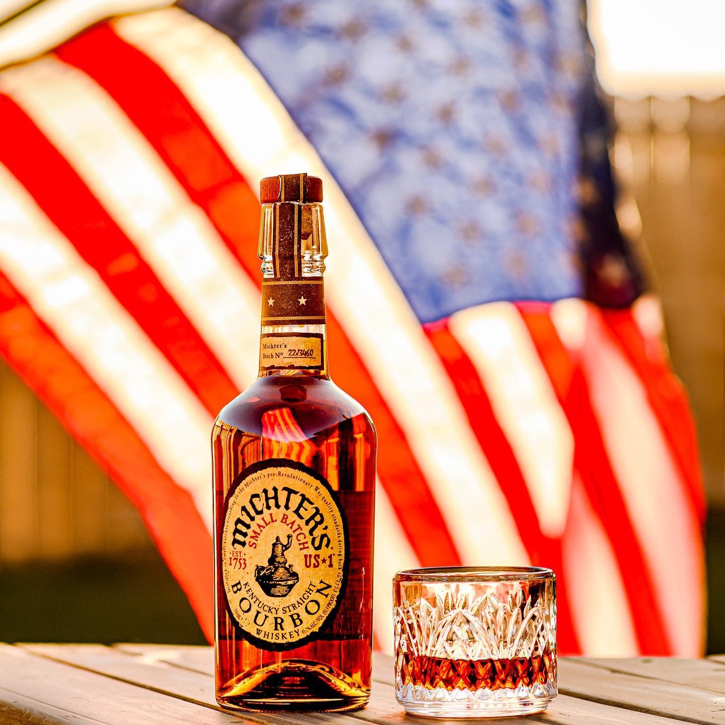 Load image into Gallery viewer, Bottle of Michter's bourbon and a glass on a wooden surface with a blurred American flag in the background.