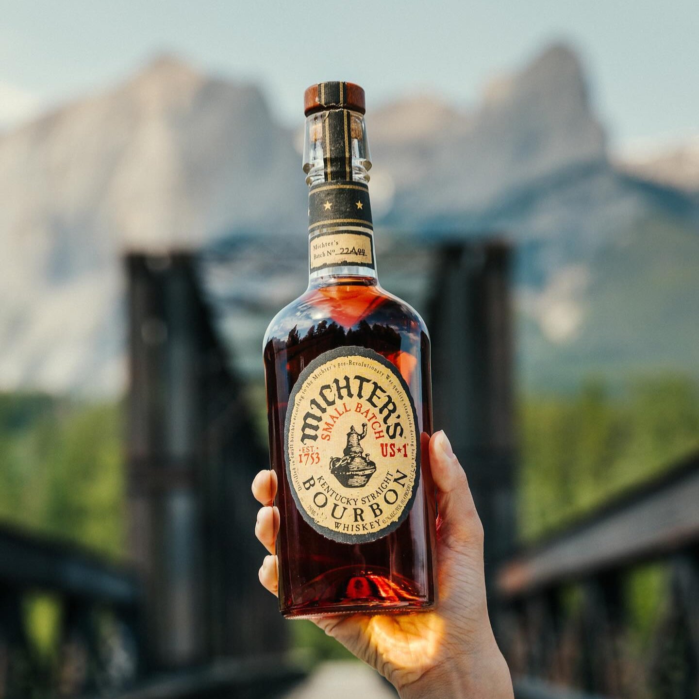 Load image into Gallery viewer, Hand holding a bottle of Michter's bourbon whiskey with a scenic mountain background.