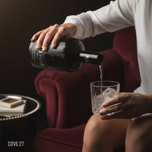 Load image into Gallery viewer, Woman in a bar pouring from a bottle of Milford Gin into a glass with ice, sitting on a plush chair.