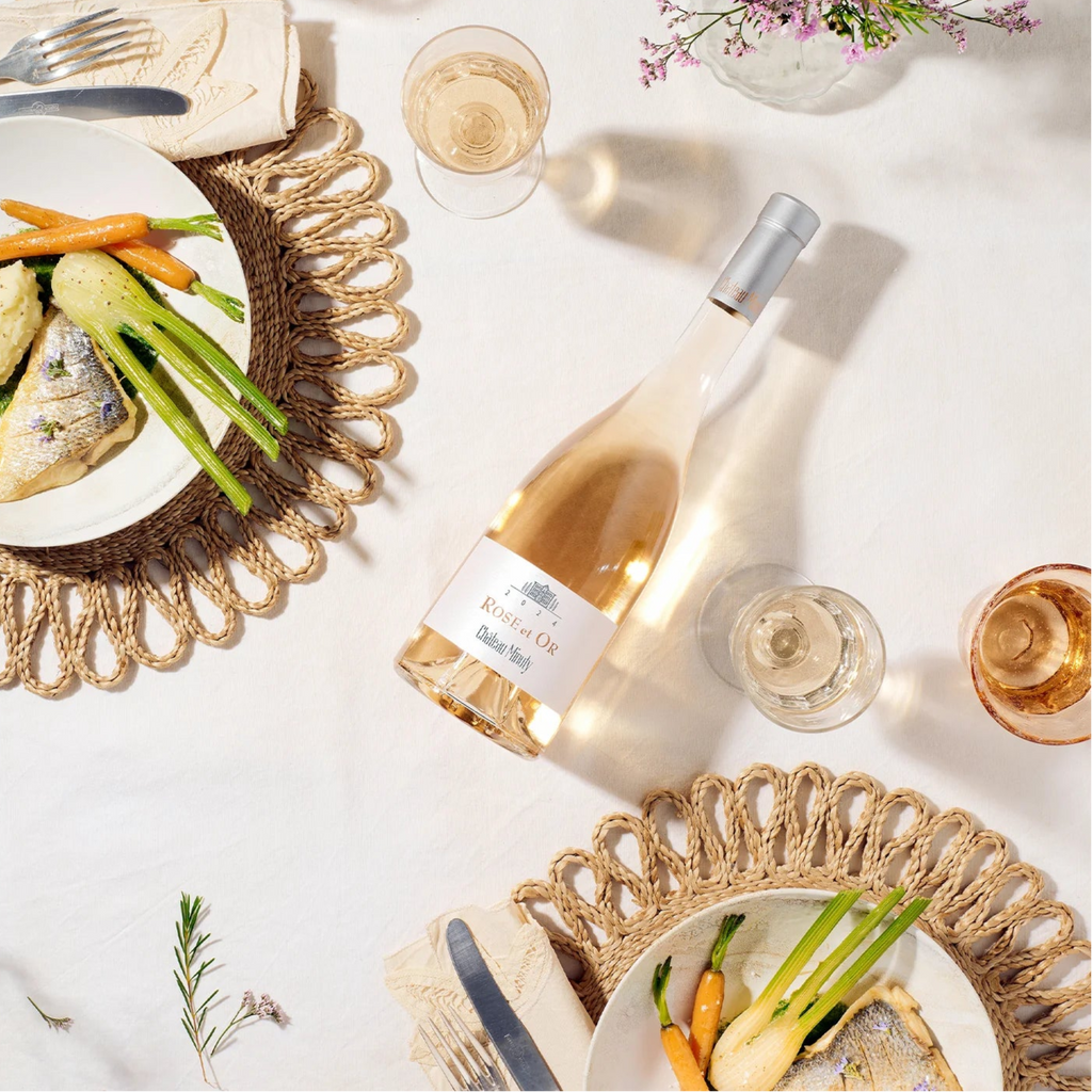 Table setting with wine bottle of Minuty Rose Et Or, plates of food, and glasses on a white surface.