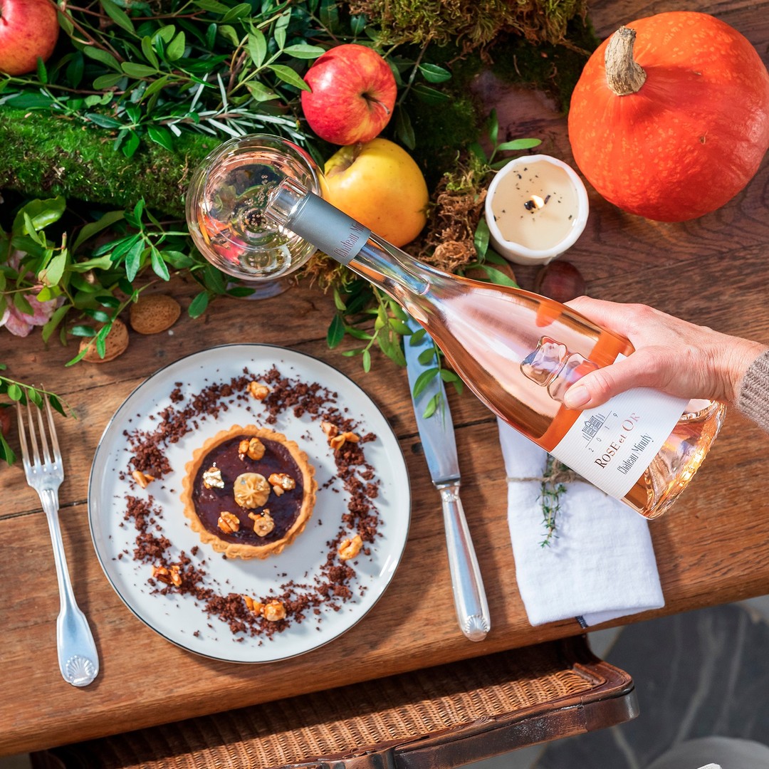 Load image into Gallery viewer, Person pouring wine into a glass on a table with a dessert, pumpkins, and apples.
