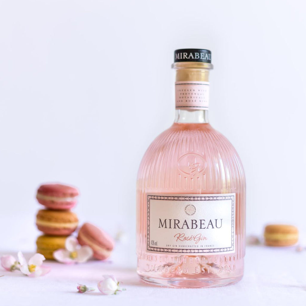 Pink bottle of Mirabeau Dry Rose Gin on a white background with macarons and flowers.
