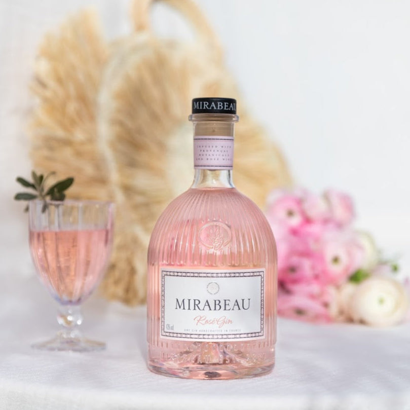Pink bottle of Mirabeau Dry Rose Gin with a glass and flowers on a light background