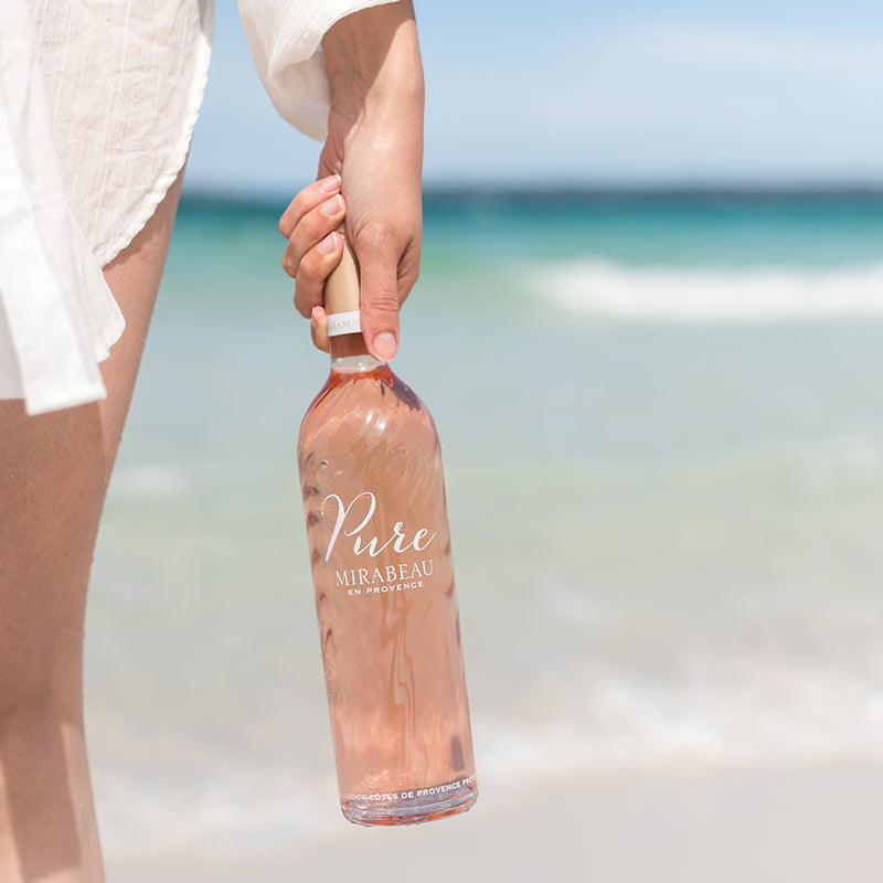 Load image into Gallery viewer, Person holding a Mirabeau Pure Provence Rosé wine bottle on a beach