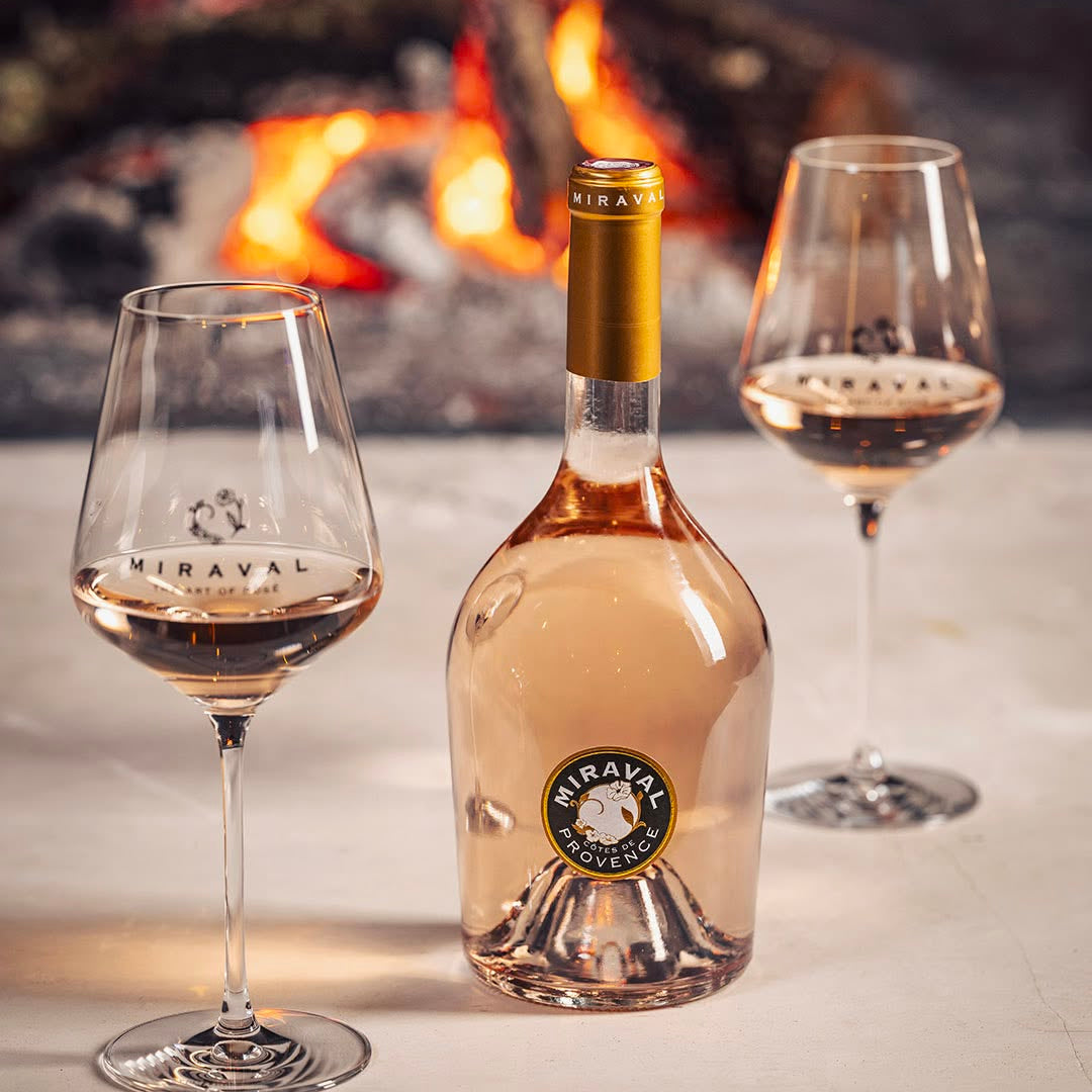 Load image into Gallery viewer, A bottle of Miraval rosé wine stands on a stone surface between two filled wine glasses, with a warm fire burning in the background.