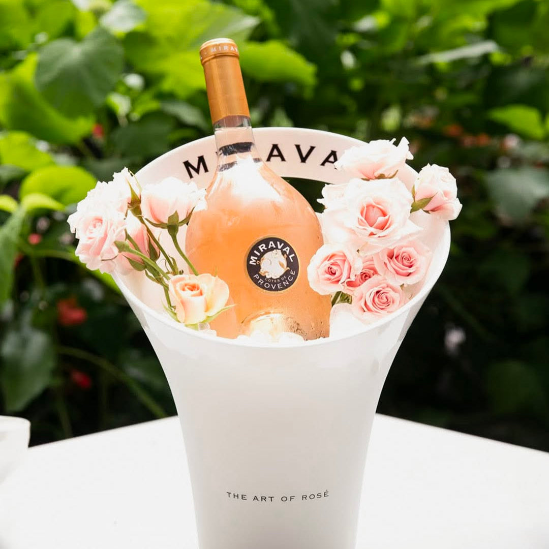 Load image into Gallery viewer, A bottle of Maraval rosé wine in a white ceramic bucket filled with ice and surrounded by pink roses, set on a white table with a blurred green garden background.