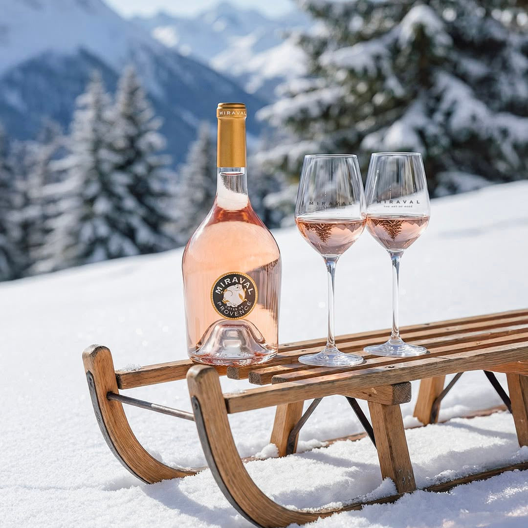Load image into Gallery viewer, A bottle of Miraval Rosé wine and two filled wine glasses placed on a wooden sled in a snowy outdoor setting with mountains and trees in the background.