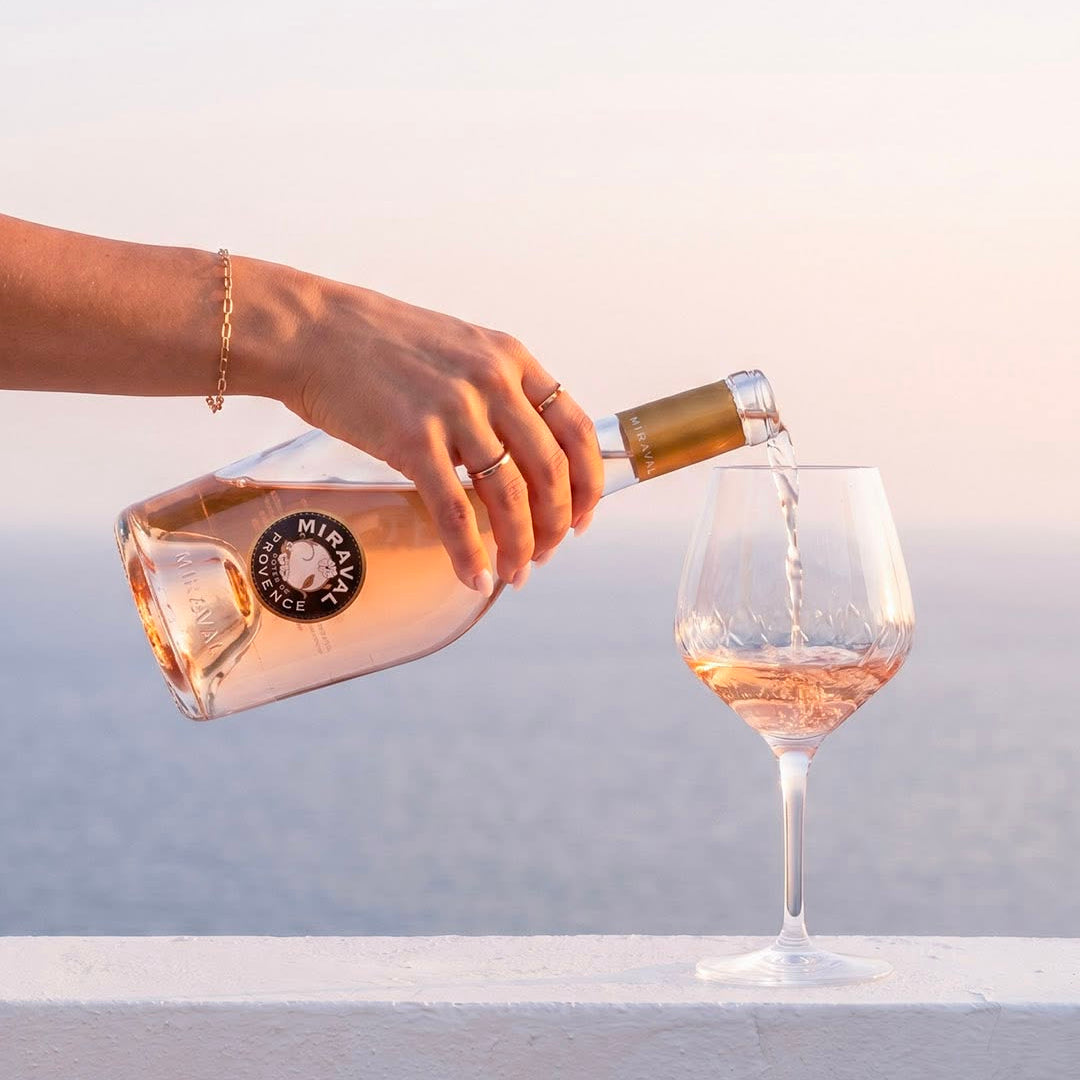 Load image into Gallery viewer, A hand pours Miraval  rosé wine into a wine glass, set against a soft, blurred coastal background at sunset.