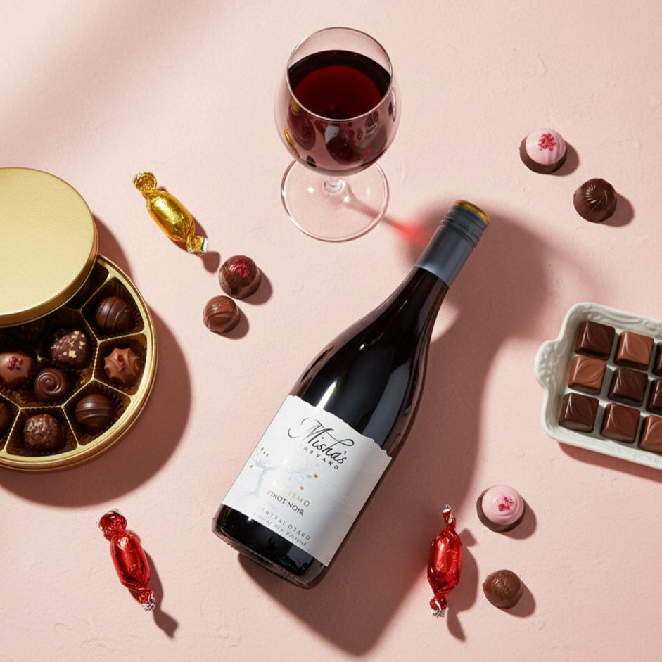 Load image into Gallery viewer, Mishas-Vineyard_Verismo_Pinot_Noir wine bottle with a glass of red wine, chocolates, and candies on a pink background