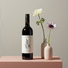 Load image into Gallery viewer, A bottle of Mitolo-Jester-Shiraz-lifestyle wine with two small vases containing flowers on a pink platform against a beige background.