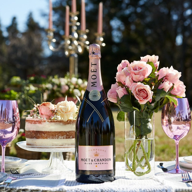 Load image into Gallery viewer, Bottle of Moet &amp; Chandon champagne on a table with pink glasses, cake, and flowers outdoors.