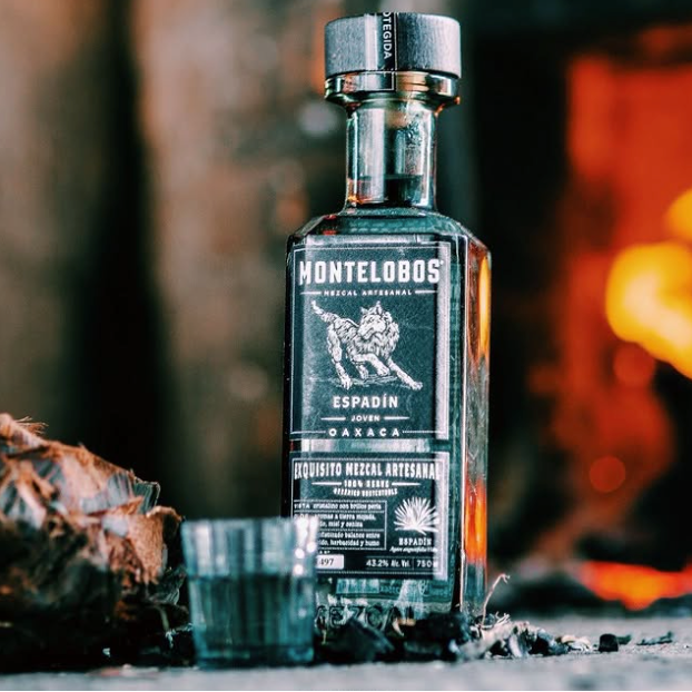Load image into Gallery viewer, A bottle of Montelobos Espadín mezcal is shown standing on a dark surface with a small matching shot glass in front. The background is a warm, blurred fire or fireplace scene, creating a rustic atmosphere.
