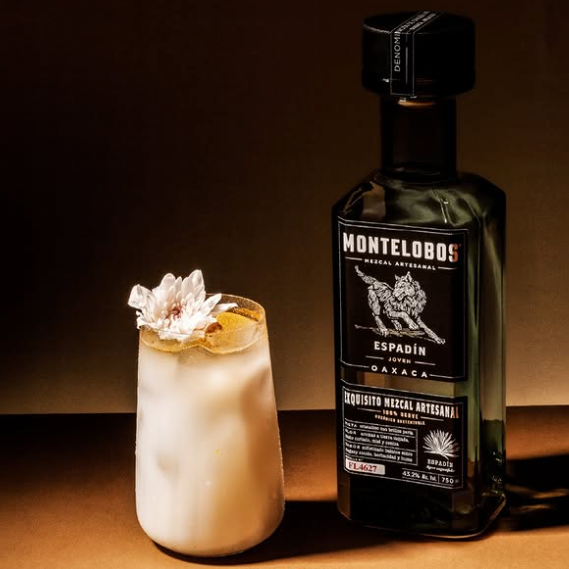 Load image into Gallery viewer, A promotional image for Montelobos Espadín Mezcal featuring a dark glass bottle next to a frothy white cocktail with a flower garnish.