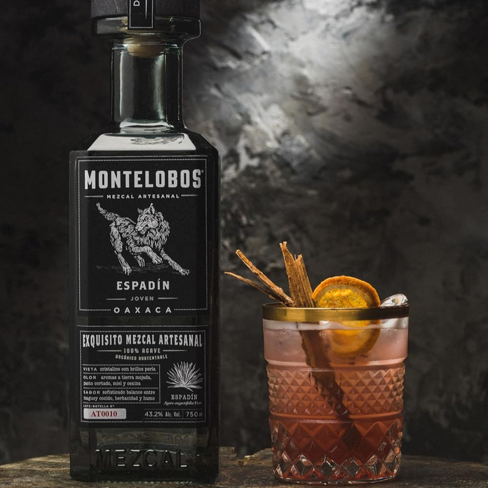 Load image into Gallery viewer, Montelobos Mezcal bottle with a cocktail garnished with an orange and cinnamon stick on a dark background