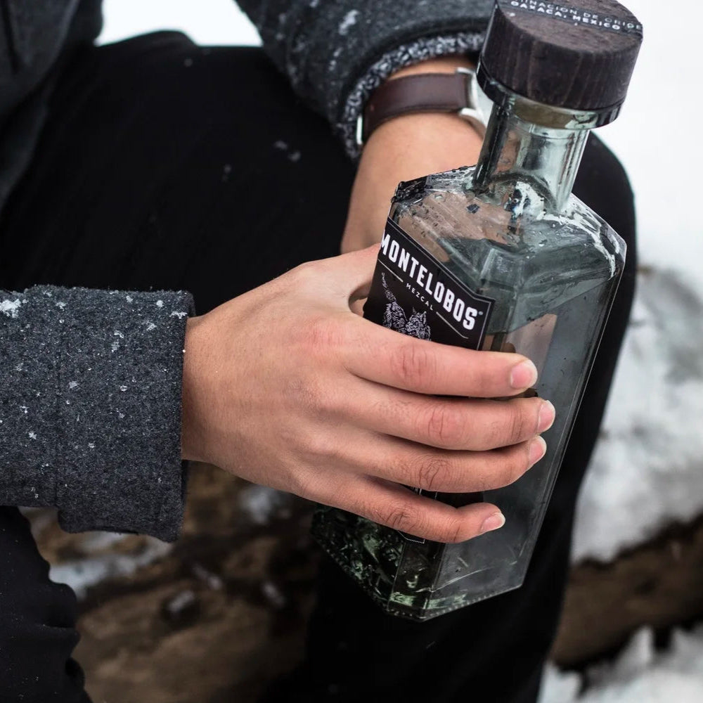 Load image into Gallery viewer, Hand holding a bottle of Montelobos mezcal in a snowy setting