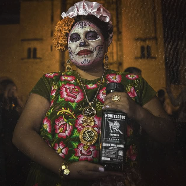 Load image into Gallery viewer, A woman in traditional Mexican Day of the Dead attire with skull face paint and a floral dress holds a bottle of Montelobos Espadin Mezcal. The background shows a dimly lit, warm-toned cathedral facade at night.