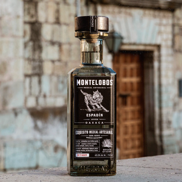 Load image into Gallery viewer, A bottle of Montelobos Espadín Oaxaca mezcal liquor stands on a stone ledge in front of a blurred rustic stone wall and wooden door.