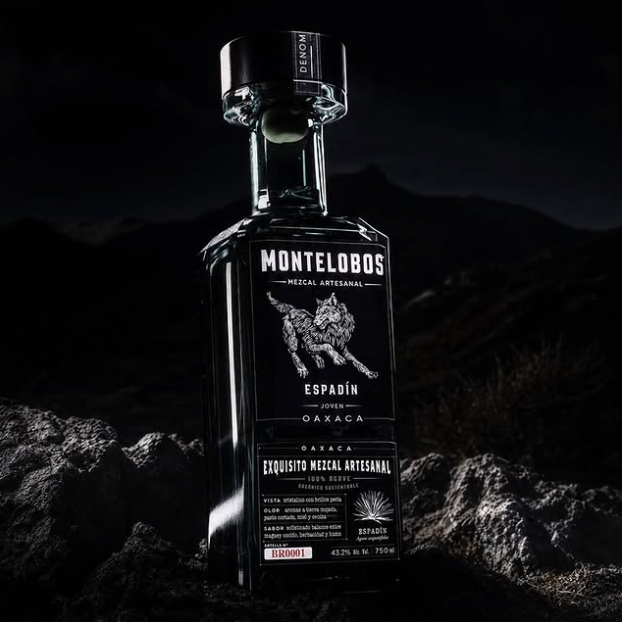 Load image into Gallery viewer, A bottle of Montelobos Espadín mezcal is photographed upright on dark rocky ground against a very dark mountainous background. The bottle is clear glass with a black cap and a black label featuring white text and a white wolf graphic.