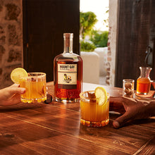 Load image into Gallery viewer, Mount Gay XO rum bottle with two glasses of a yellowish-orange cocktail on a wooden table.