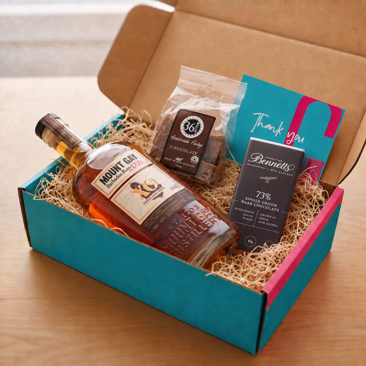 Load image into Gallery viewer, A curated gift box set on a wooden table, featuring a bottle of Mount Gay XO rum, a packet of 36 Degrees chocolate fudge, a bar of Bennett's 73% Single Origin Bar Chocolate, and a teal and pink COVE 27 thank you card, all arranged in a turquoise and pink COVE 27 box filled with wood-wool.