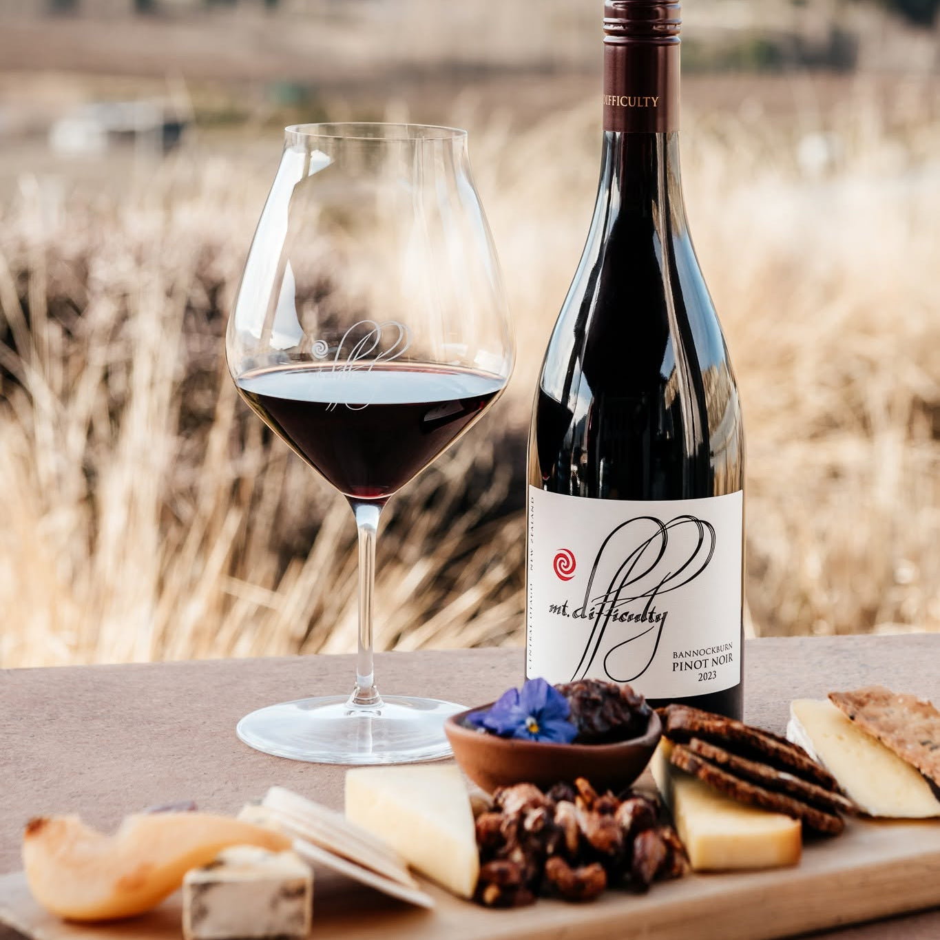 Load image into Gallery viewer, A bottle of Mt Difficulty Bannockburn Pinot Noir red wine and a filled wine glass on a wooden board with cheese, crackers, and nuts, set outdoors against a blurred field background.