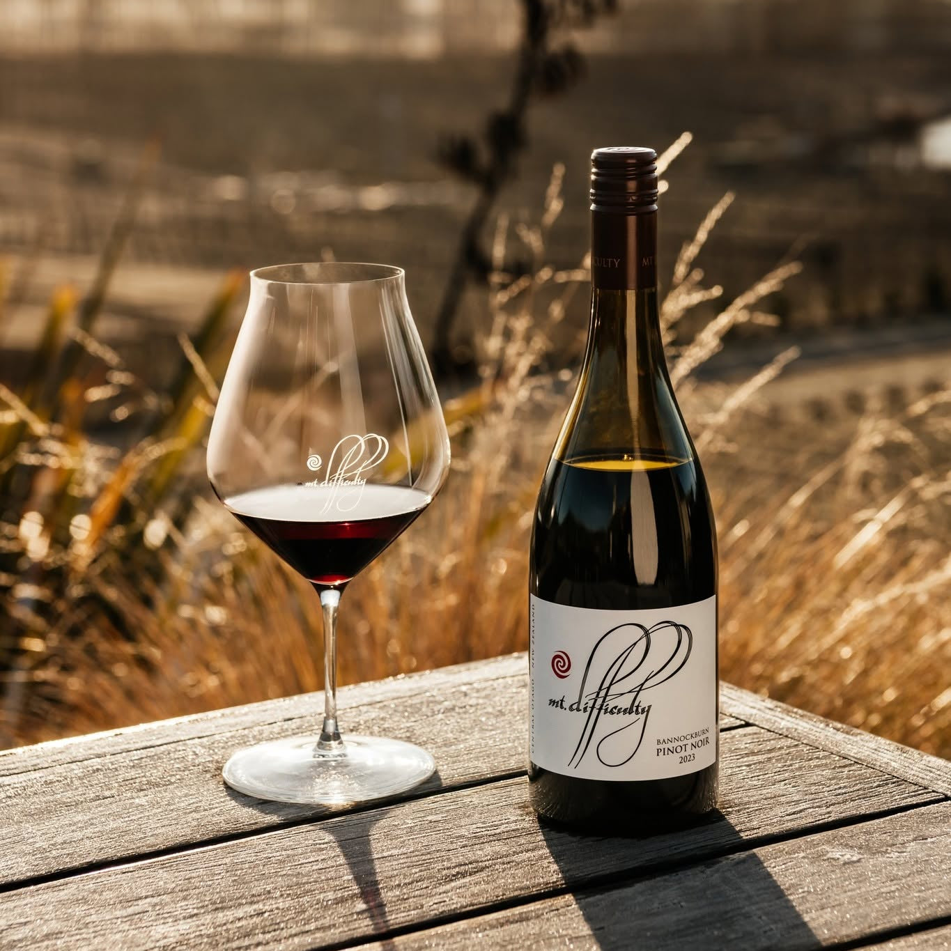 Load image into Gallery viewer, A bottle of Mt Difficulty Bannockburn Pinot Noir red wine and a stemmed glass filled with red wine sit on a weathered wooden table outdoors, with dry grass and a softly blurred landscape in the background.