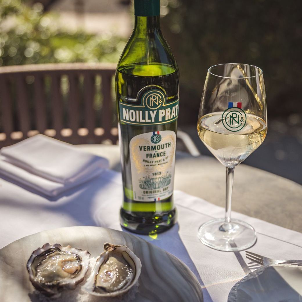 Load image into Gallery viewer, A bottle of Noilly Prat Original Dry Vermouth and a glass and some oysters on a plate on a table with some greenery in the background