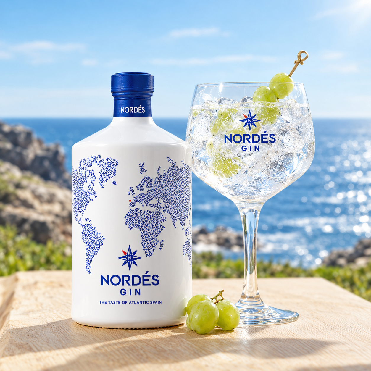 Load image into Gallery viewer, A bottle of Nordés Gin and a filled coupe glass with ice, green grapes, and a grape skewer, set on a wooden board with a sunny ocean background.