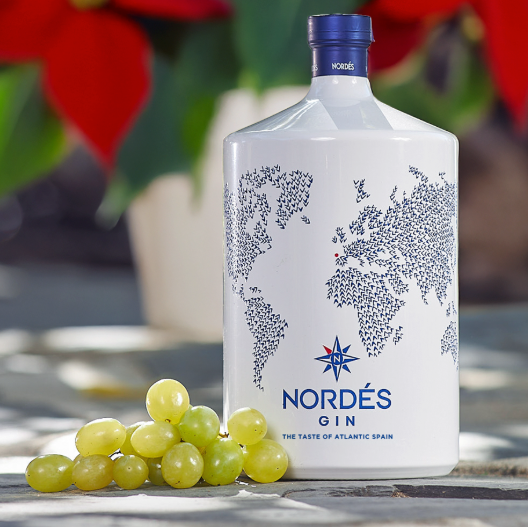 Load image into Gallery viewer, A bottle of Nordés Gin gin is displayed on a stone surface with a small pile of green grapes in the foreground and blurred red flowers in the background.