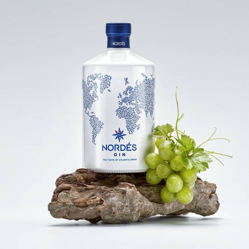 Load image into Gallery viewer, A bottle of Nordés Gin on a piece of driftwood next to a bunch of green grapes, set against a light gray studio background.