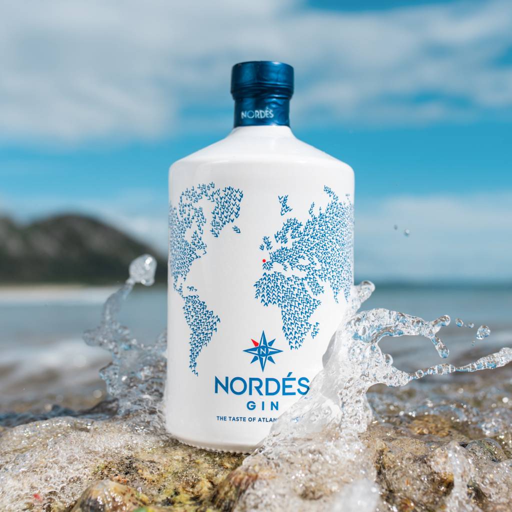 Load image into Gallery viewer, A bottle of Nordés Gin stands on rocks with ocean water splashing around it, against a blurred beach and sky background. The bottle is white with a blue world map graphic and blue branding text.