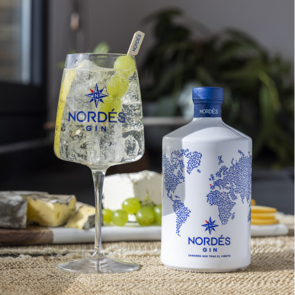 Load image into Gallery viewer, A bottle of Nordés Gin stands beside a tall glass filled with a clear cocktail, garnished with a lime wheel and a straw, set on a burlap surface with cheese, grapes, and a blurred indoor background.