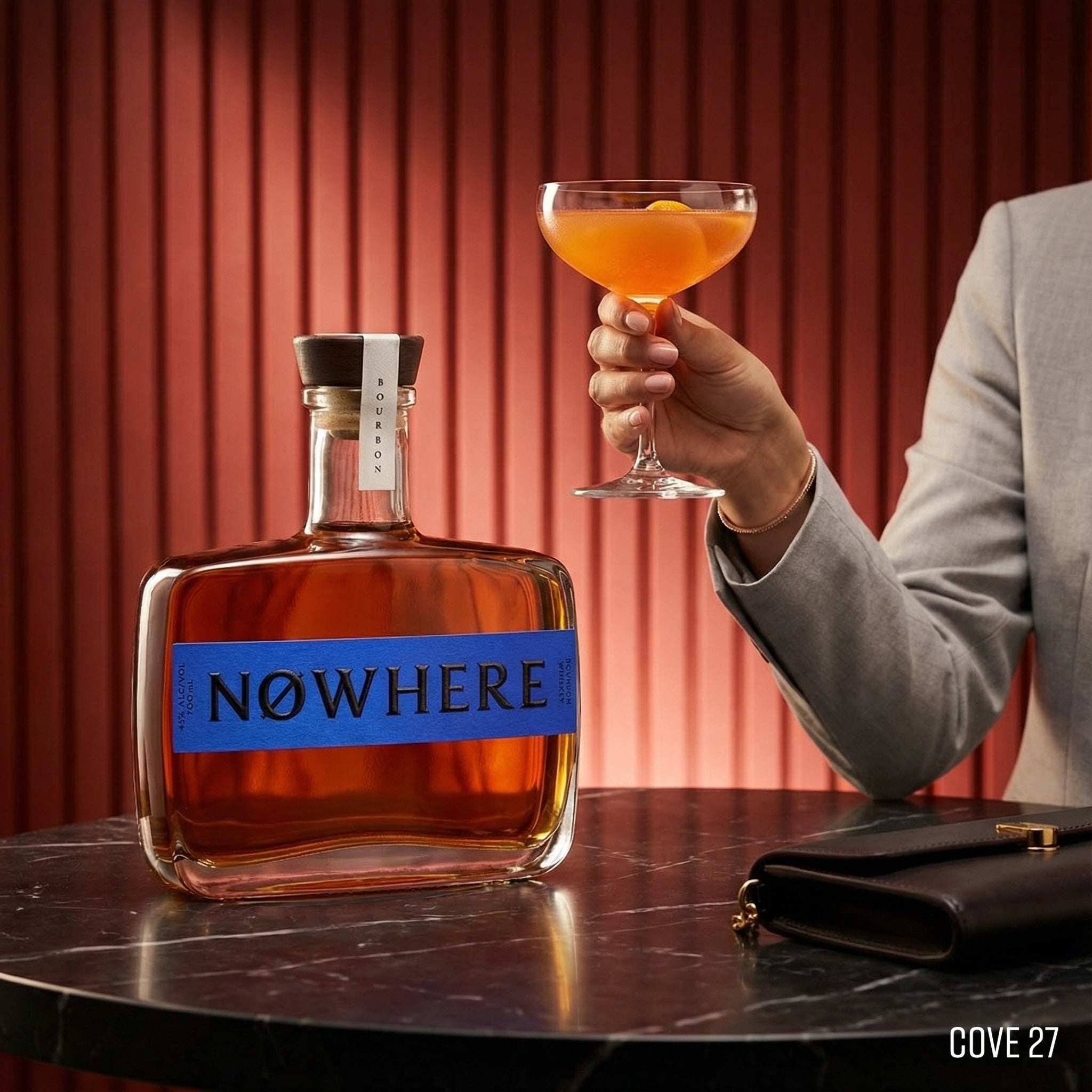 Load image into Gallery viewer, Bottle of 'NOWHERE' whiskey with a hand holding a cocktail against a red curtain background.