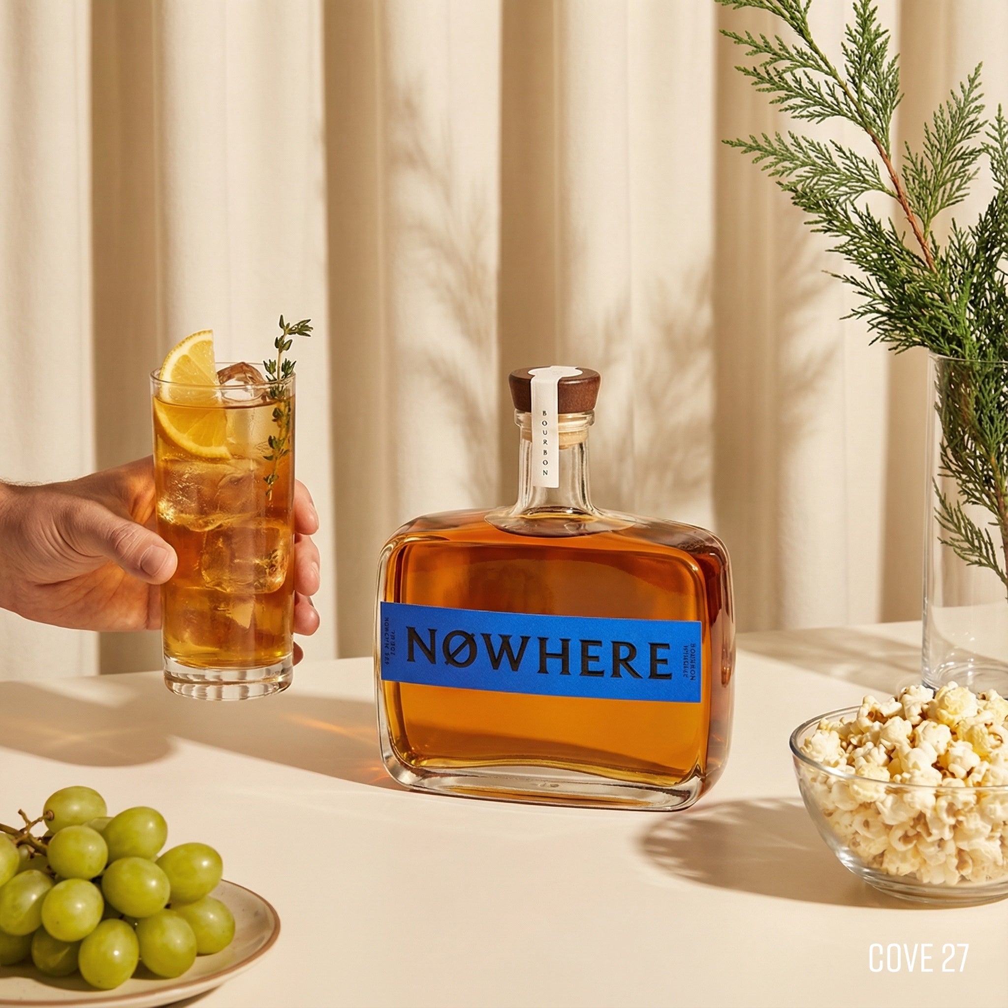 Load image into Gallery viewer, Bottle of 'NOWHERE' whiskey with a glass of iced tea, grapes, and popcorn on a table.