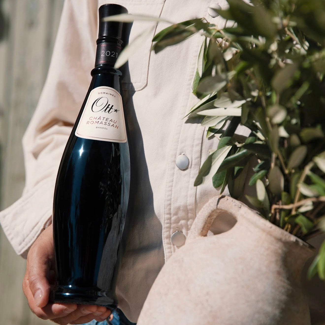Load image into Gallery viewer, A person in a light beige shirt holds a dark bottle of Chateau Romassan Bandol red wine, with a potted olive plant in the foreground.